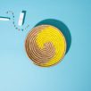 Handgemachte Schale aus Sisal und Süssgras aus Ruanda, mit einem spiralförmigen Muster in den Farben Gelb und Beige auf einem blauen Hintergrund. Neben der Schale liegen ein Lippenstift, ein kleines Parfümfläschchen und ein buntes Armband.