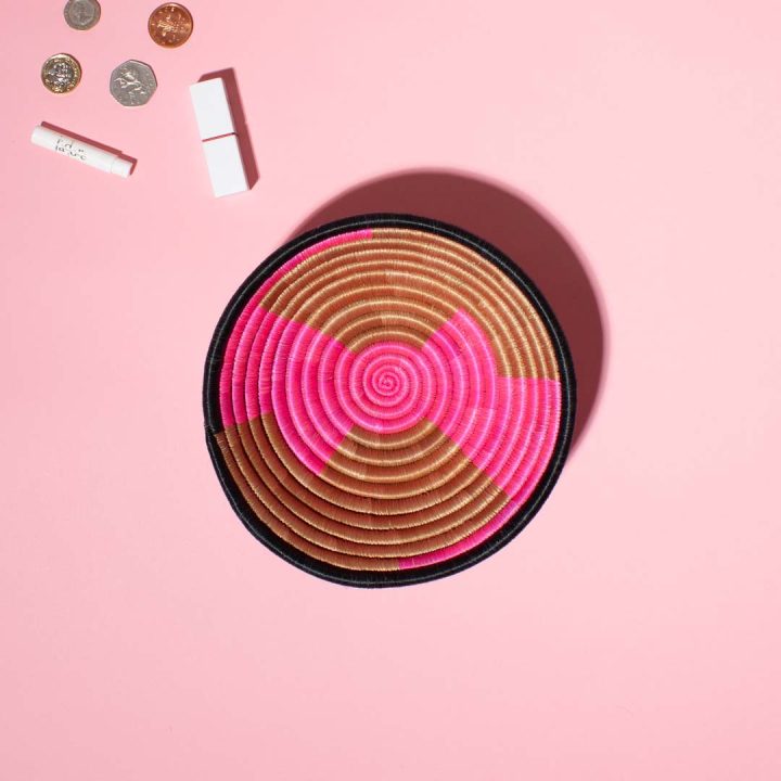 Eine handgemachte runde Schale aus Sisal und Süssgras, hergestellt in Ruanda, auf einem rosa Hintergrund. Der Korb hat ein geometrisches Muster in leuchtendem Pink und Beige, mit einem schwarzen Rand. Links oben sind einige Münzen, ein Lippenstift und ein kleines Parfümfläschchen arrangiert.