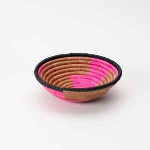 Seitenansicht einer handgemachten runden Schale aus Sisal und Süssgras, hergestellt in Ruanda, auf einem weissen Hintergrund. Der Korb hat ein geometrisches Muster in leuchtendem Pink und Beige, mit einem schwarzen Rand.